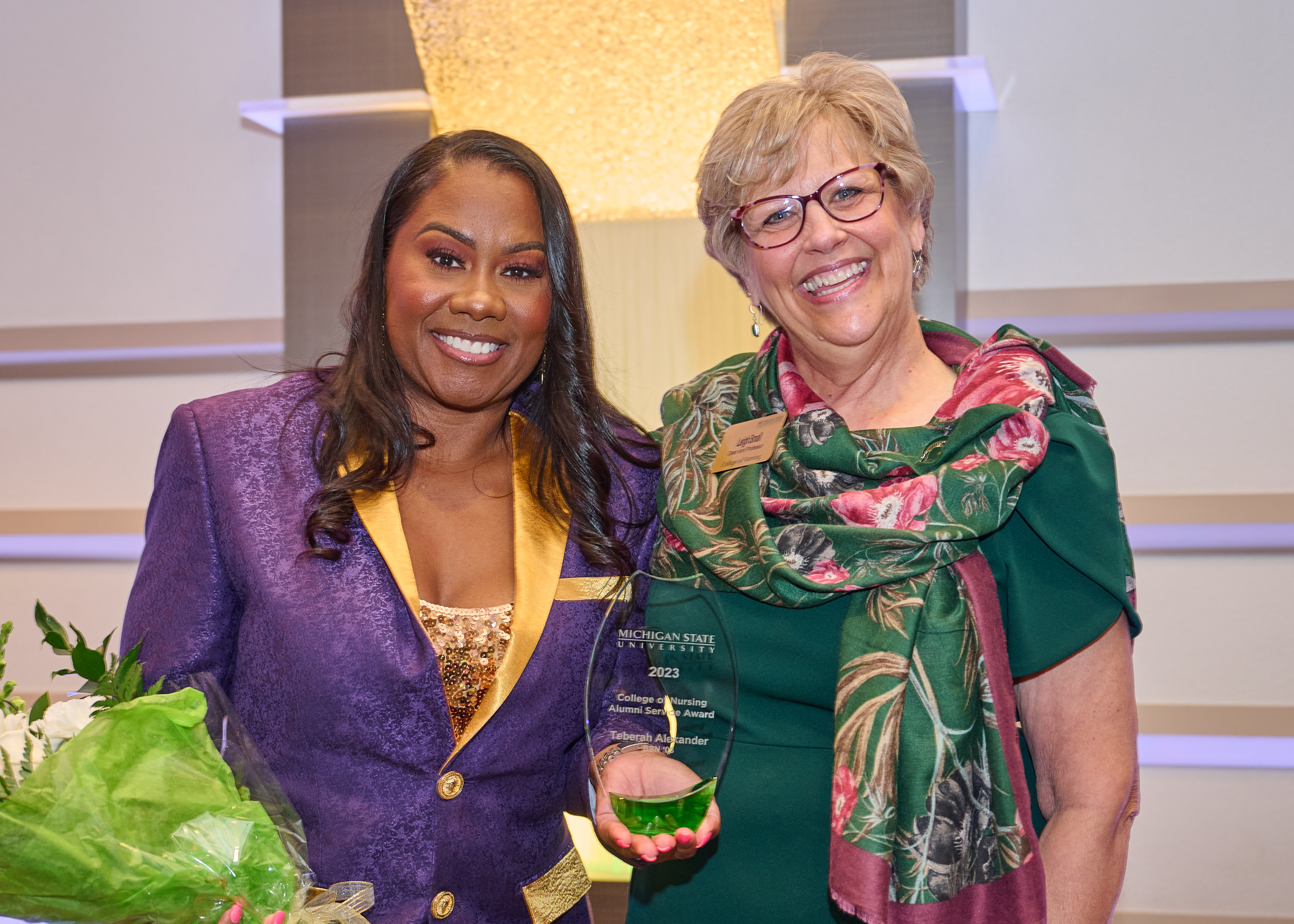Teberah Alexander holds a flower bouquet and a crystal trophy for winning the Alumni Service Award. She smiles for a photo along with Dean Leigh Small.