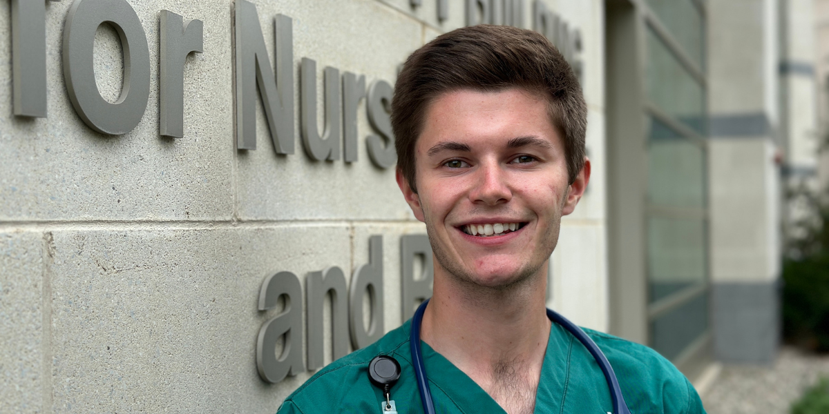 Caleb Fraser smiling at the camera in scrubs leaning up against a cinderblock building 