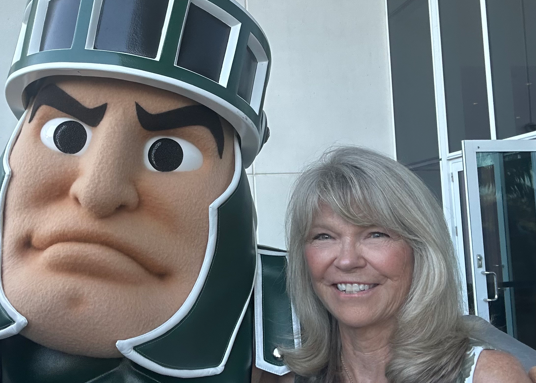 Nancy King Reame, a College of Nursing alumna, takes a selfie with Sparty, MSU's mascot, in front of a stone building.