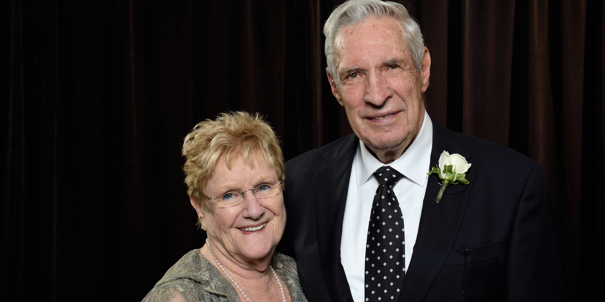 A couple poses together in formal attire for a portrait. The individual on the right is wearing a black suit with a white shirt, a black polka-dot tie, and a white boutonnière. The individual on the left is wearing a lace-patterned dress with a necklace. Both are smiling in front of a dark backdrop.