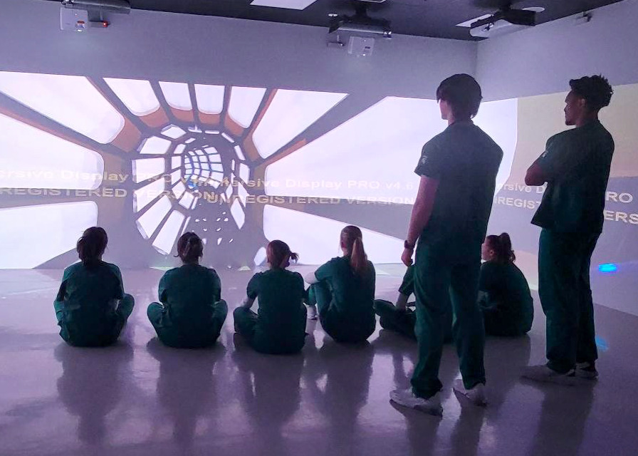 Several current College of Nursing students watch a fully-immersive video of a space ship as they experience with a simulation room.
