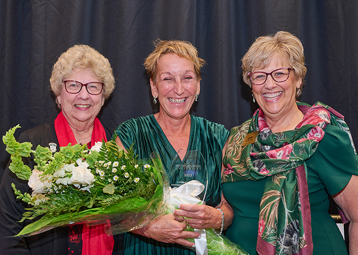 Joan Brueggeman stands in between Angie Strawn and Dean Leigh Small while they pose for a photo. All three people are smiling, and Joan holds a crystal trophy and flower bouquet after winning the Distinguished Alumni Award.