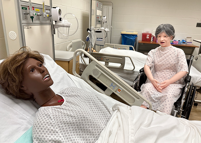 Two College of Nursing mannekins rest in the CON simulation lab, waiting for students to come in. One mannekin is set up on a hospital bed, and the other is set up in a wheelchair.