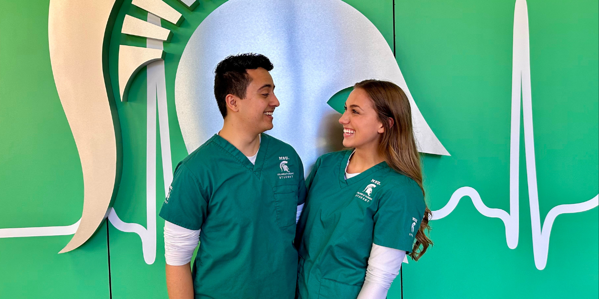 Two students looking at each other and smiling in scrubs. They are a man and a woman. 