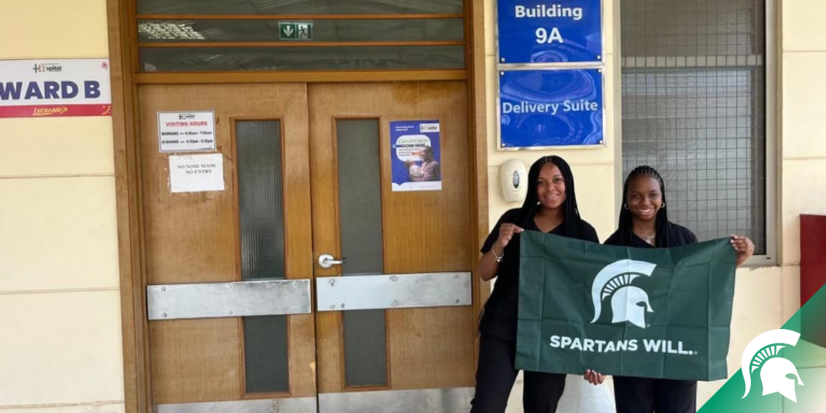 Two students holding a 'Spartans Will' banner proudly stand outside a hospital building labeled 'Ward B' and 'Building 9A: Delivery Suite.' The students are smiling, showcasing their school spirit in a professional healthcare setting, emphasizing their affiliation with Michigan State University. The background displays the hospital's entrance doors and informational signs about visiting hours and entry restrictions