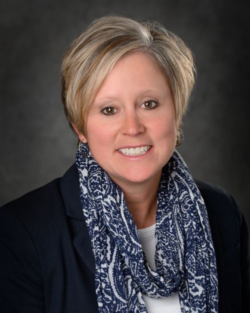 Photo of Patty West has short blond hair framing her face, wears a navy blazer and white shirt under it, with a navy blue and white scarf smiles at camera