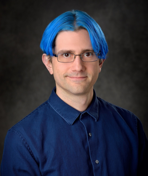 Photo of Robert Crawley smiling at camera has short blue hair and glasses on a portrait backdrop.